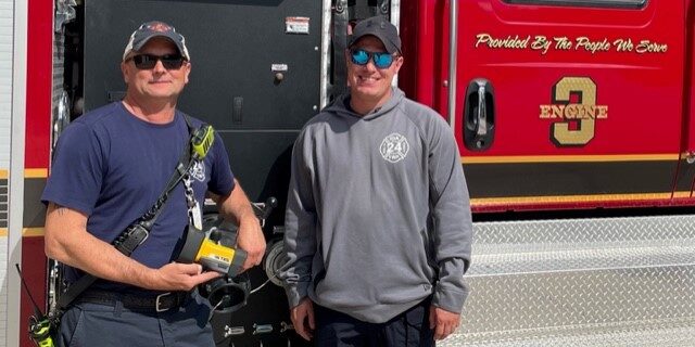 Two male firefighters stand in front of a fire engine holding a thermal imaging camera.