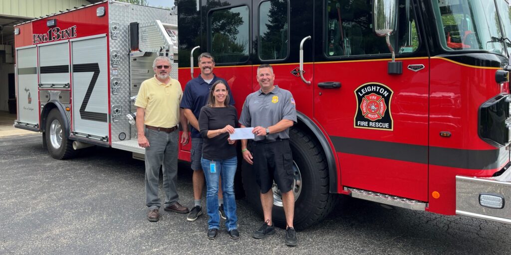 A man and woman from MGU present two male firefighters with a grant check next to a fire engine.