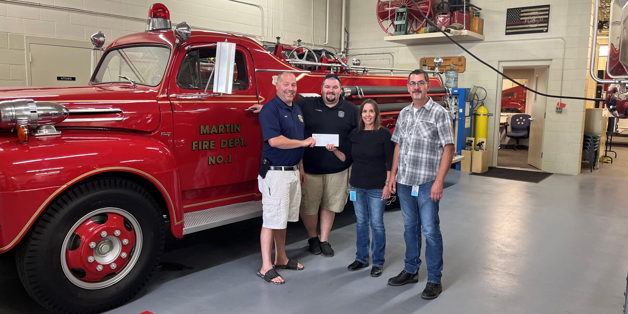 Two male firefighters receive a grant check from a man and woman with MGU in front of a fire engine.