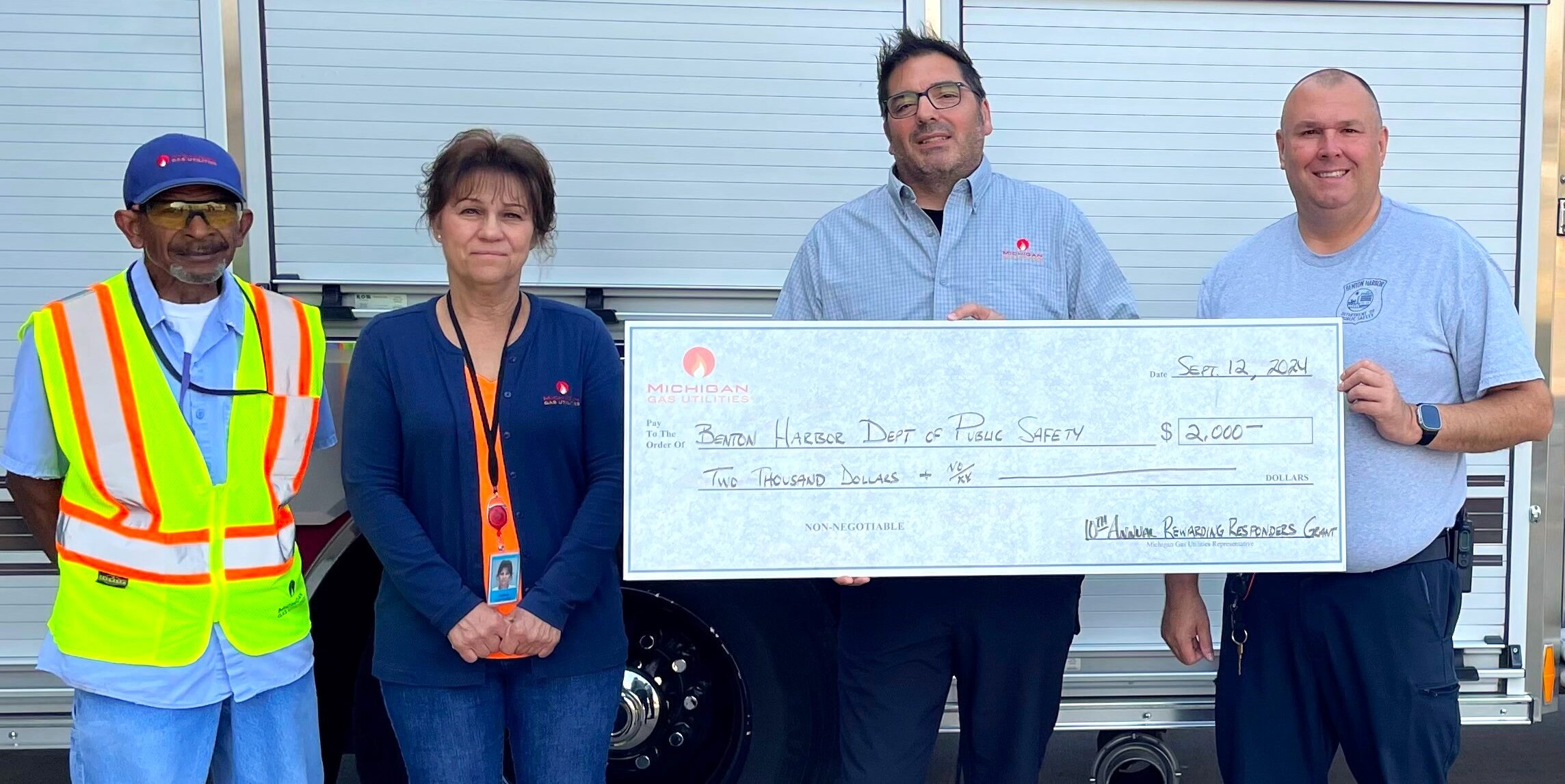 Two men hold a large ceremonial check next to another man and a woman. The group stands in front of a fire engine.