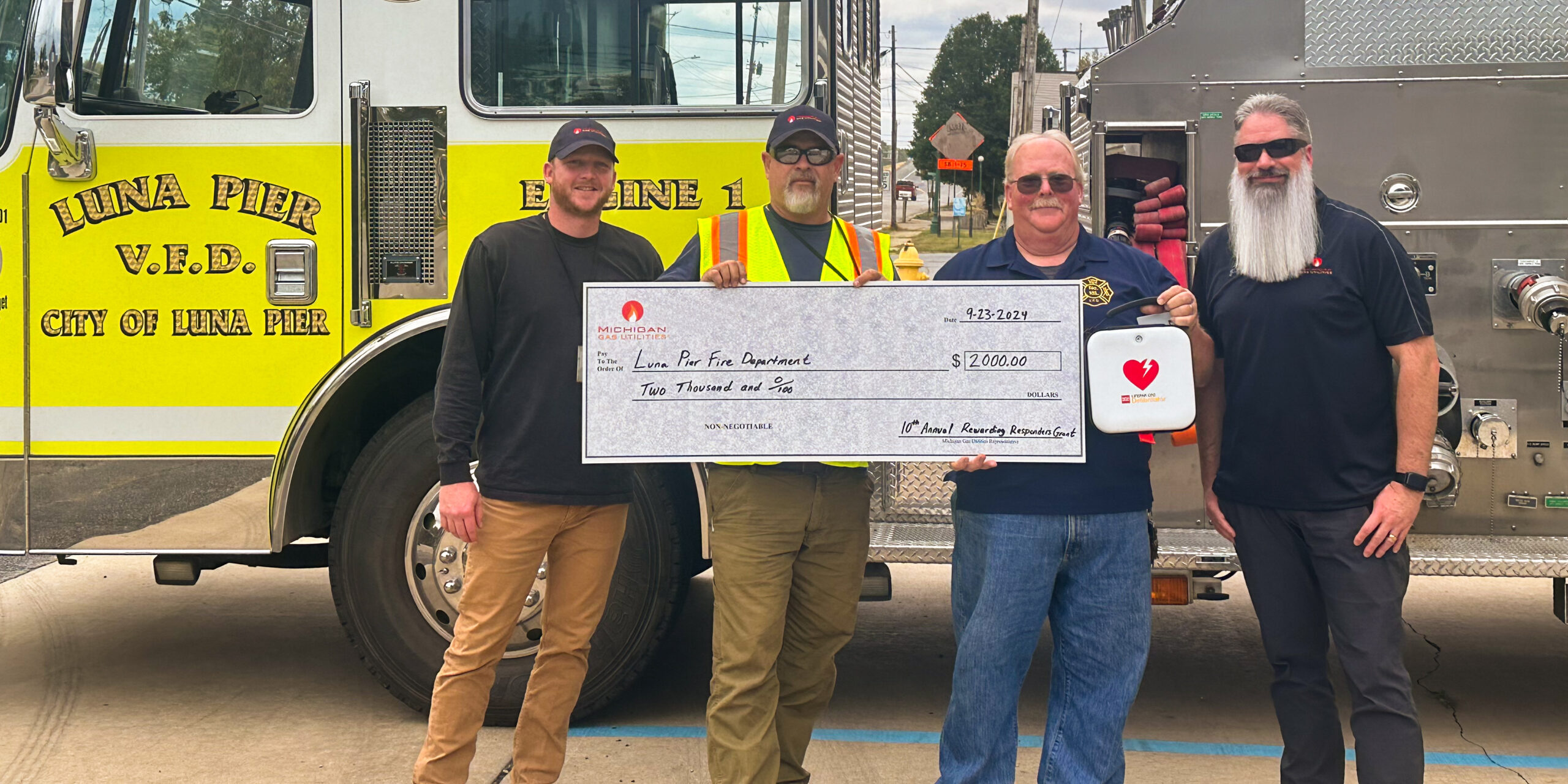 A group of male MGU employees hold a large ceremonial check with the Luna Pier Fire Department fire chief in front of a fire engine.