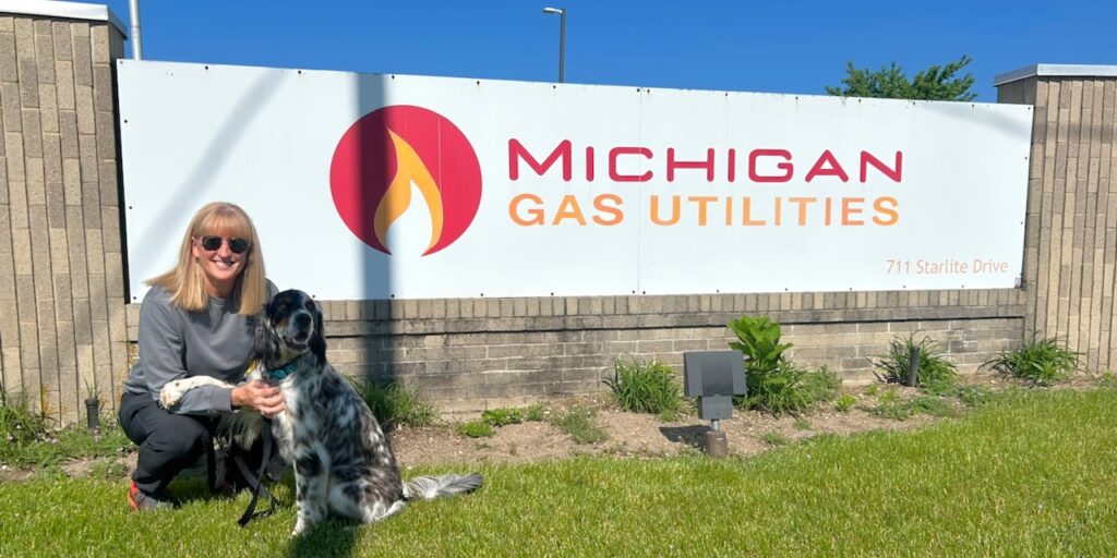 A woman kneels next to a dog with black and white hair in front of a rectangular sign with the MGU logo on it.