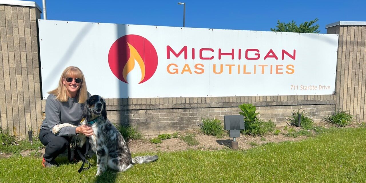 A woman kneels next to a dog with black and white hair in front of a rectangular sign with the MGU logo on it.