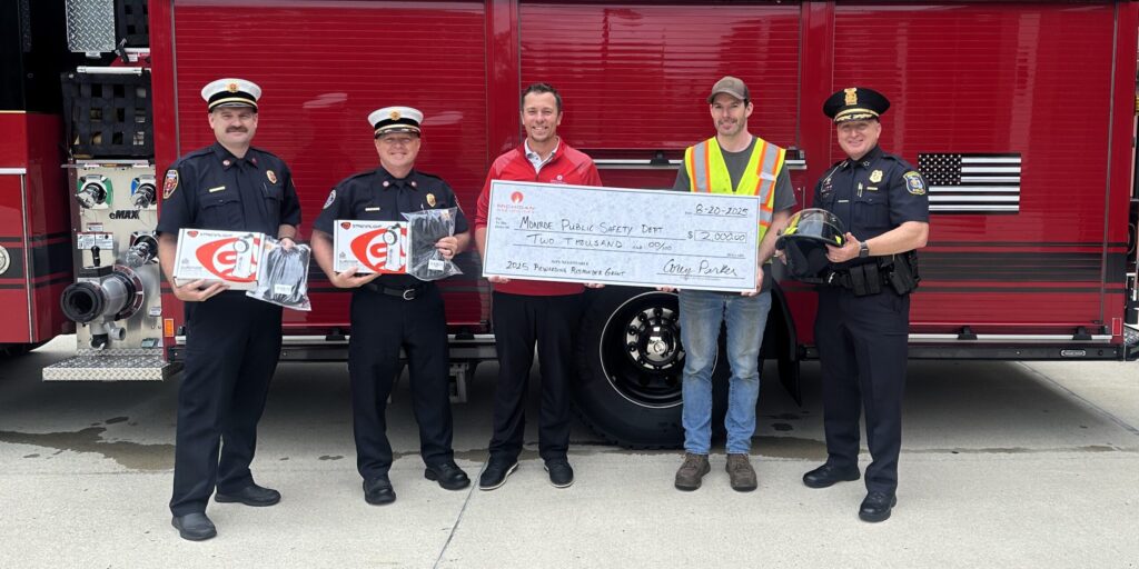 Two men from MGU hold up a large ceremonial check surrounded by firefighters holding up protective clothing in front of a fire engine.