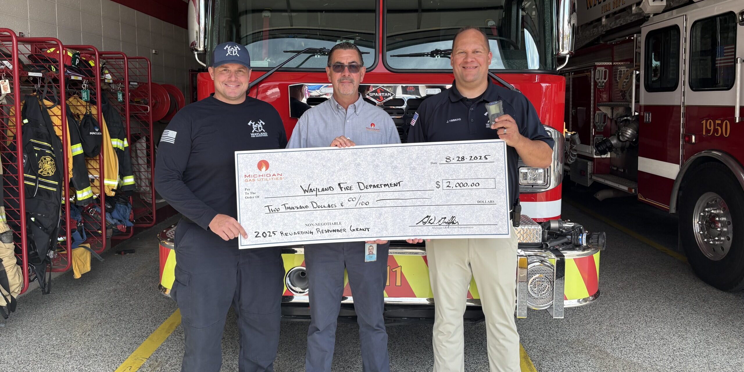 A male MGU employee holds up a large ceremonial check while standing next to two firefighters from the Wayland Fire Department.