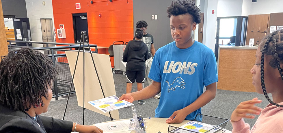 A teenage boy shares project materials with two teenage girls inside a Boys and Girls Club facility.