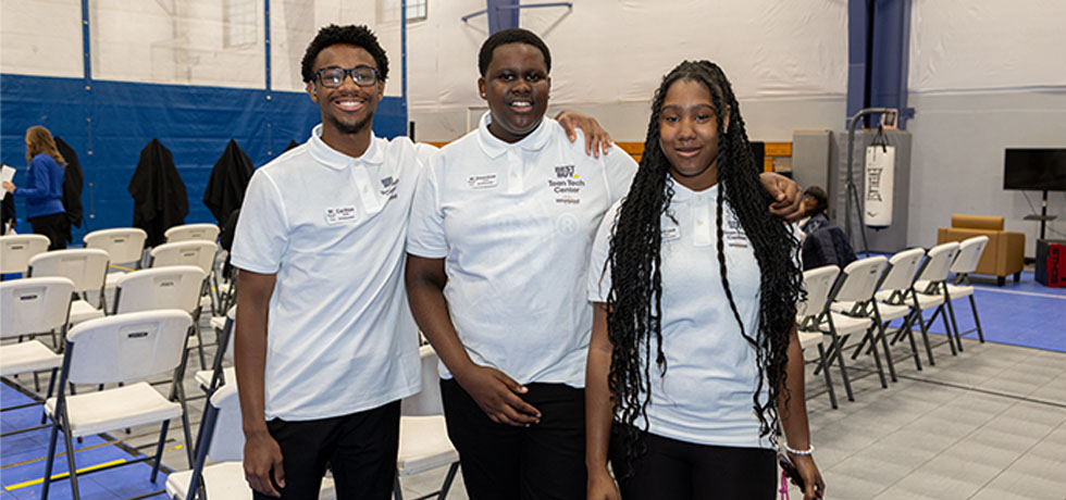 Two teenage boys and a teenage girl smile for a photo inside a multipurpose room.
