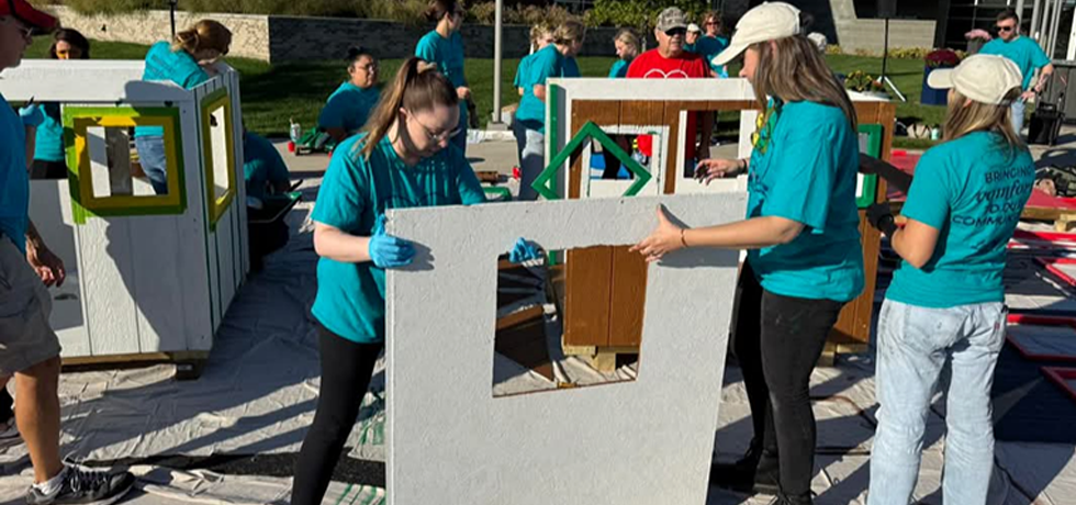 A group of men and women help build small house structures on a sunny day.