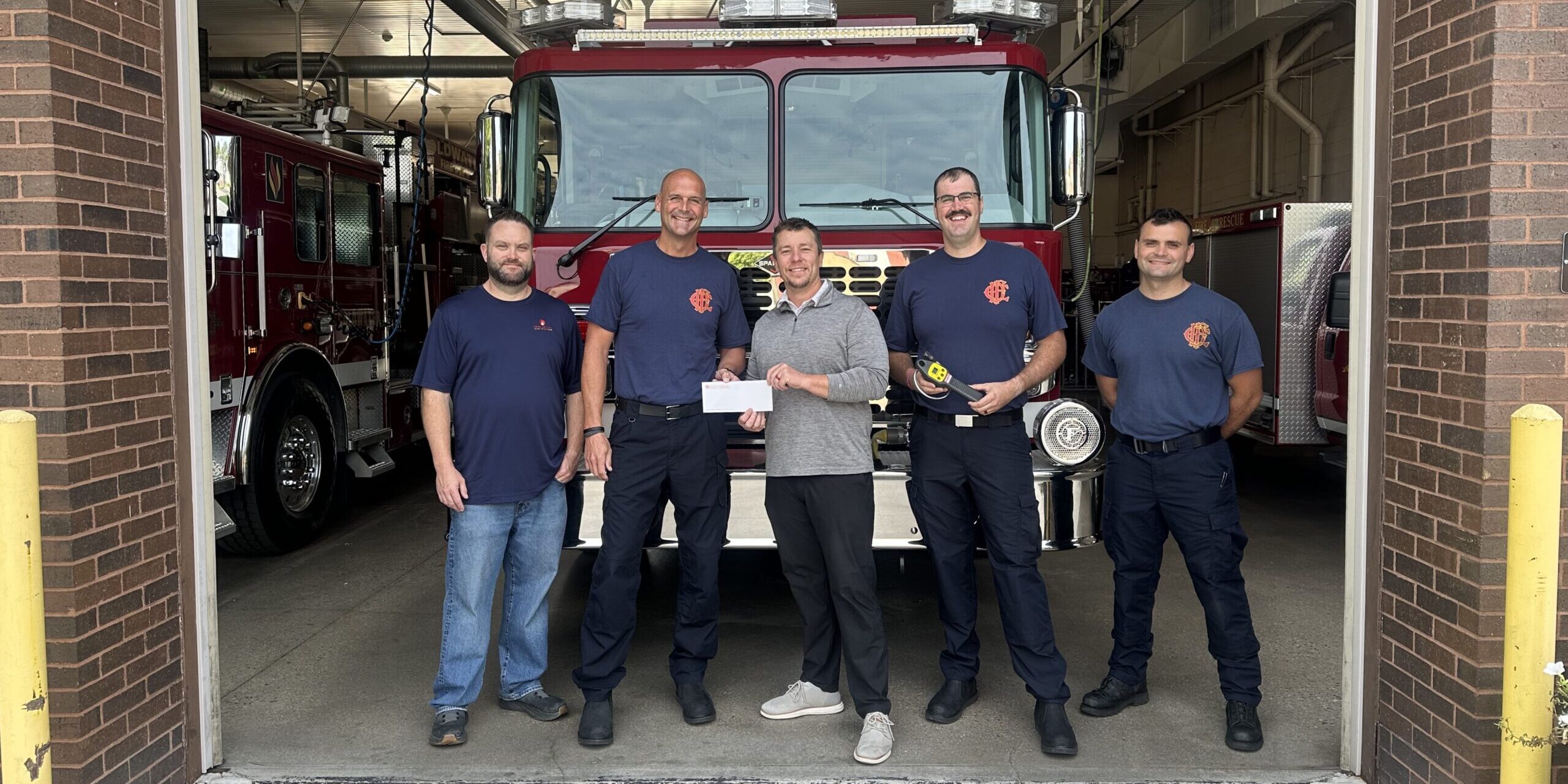 A man with MGU presents a donation check to firefighters with the Coldwater Fire Department while standing in front of a fire truck in a garage.
