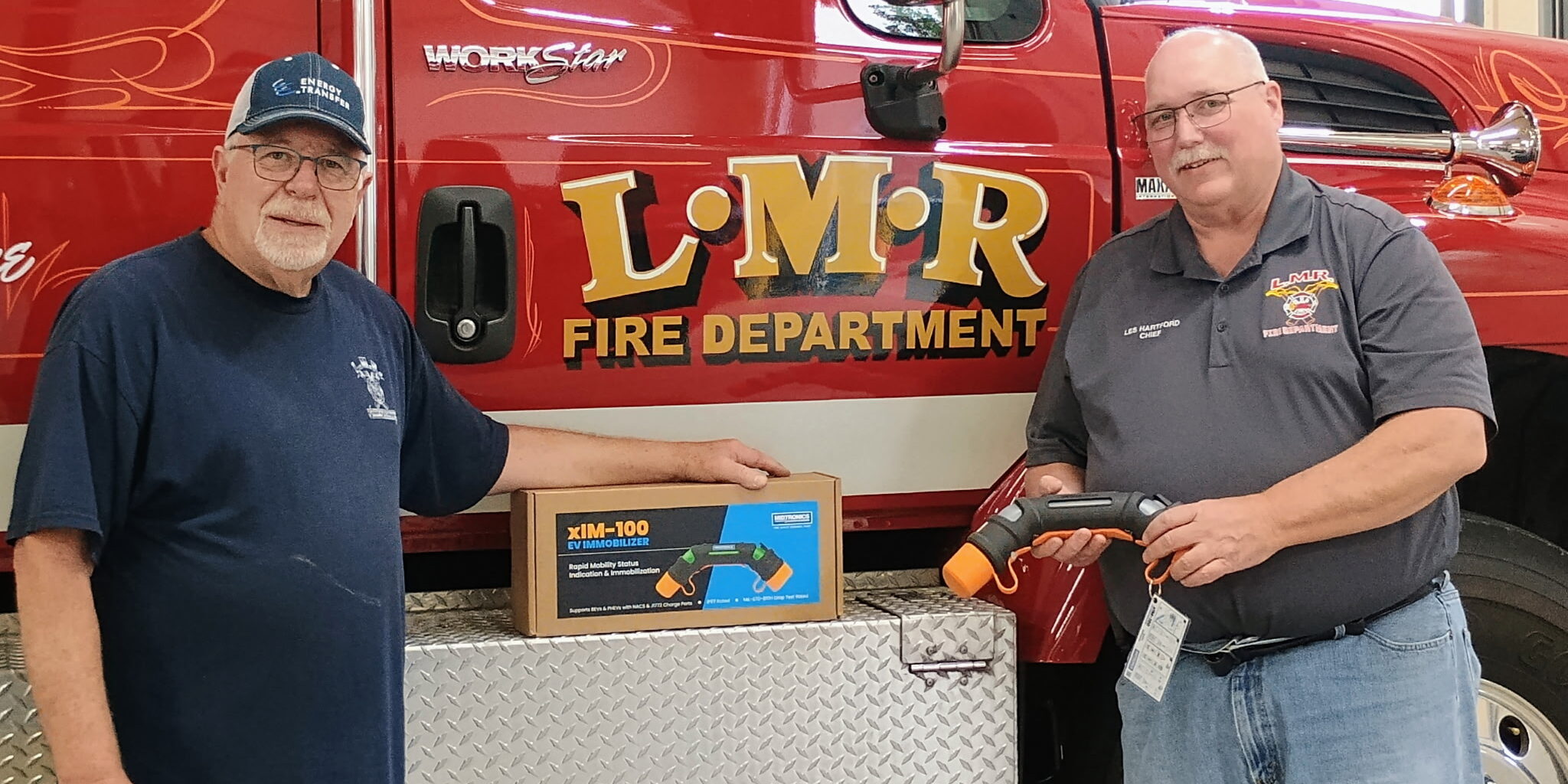 Two men take a photo with an electric vehicle immobilizing device while standing in front of a fire truck.