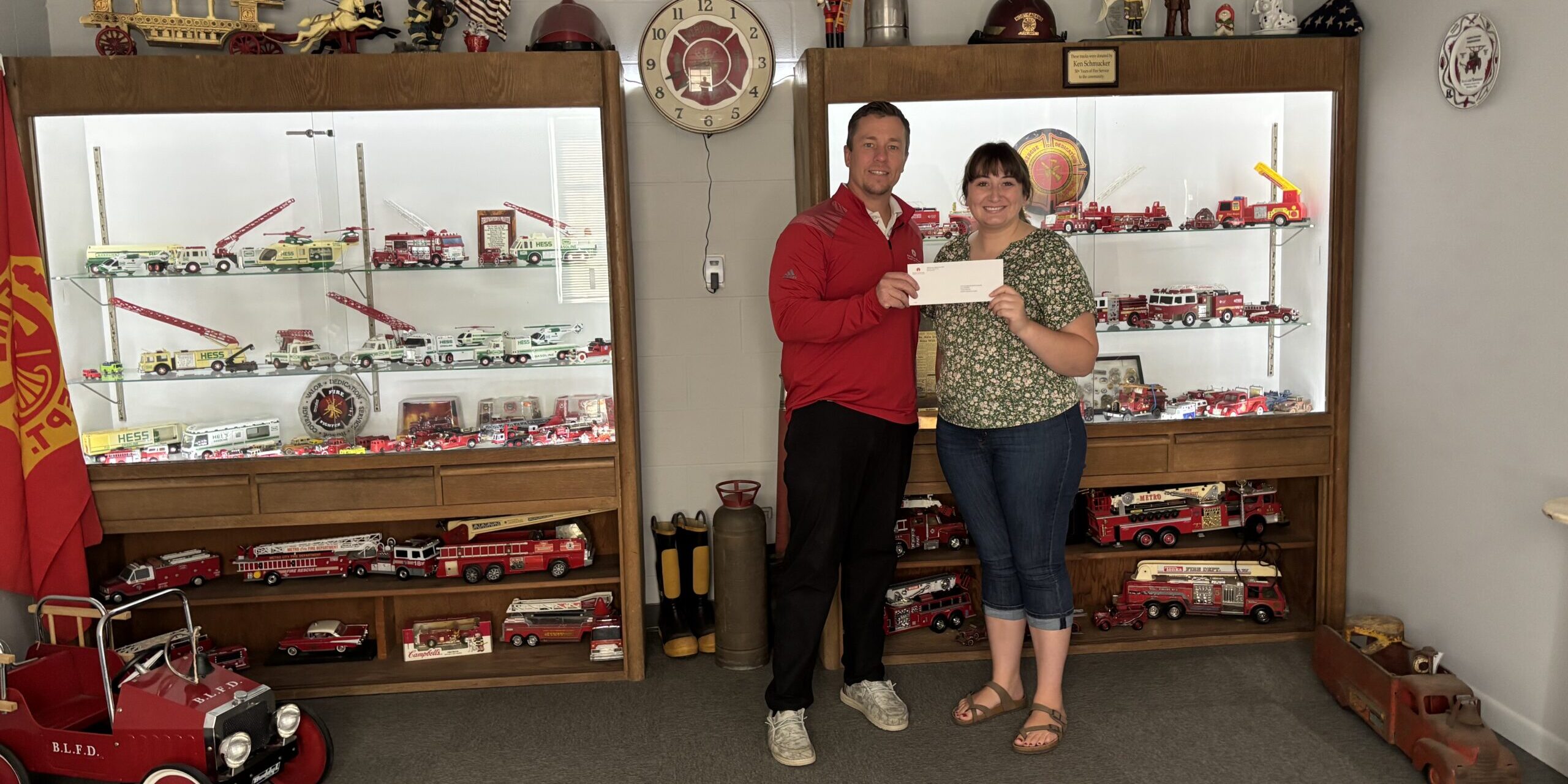 A man with MGU and a woman from the North Adams Fire Department take a photo holding a donation check in a room of fire department memorabilia.