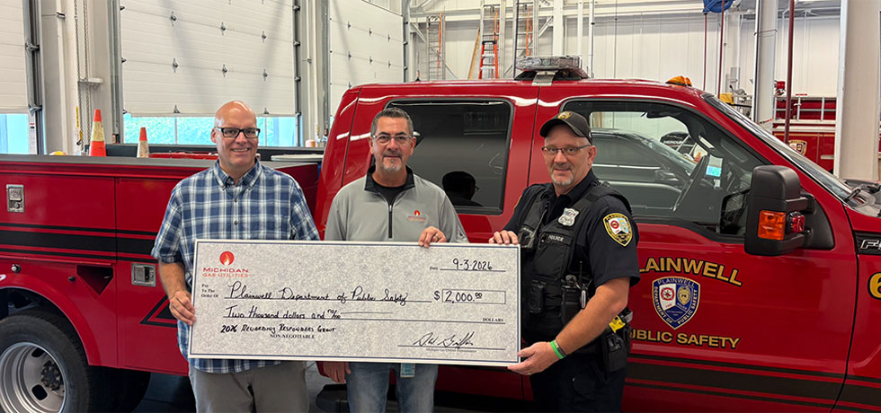 Two men with the Plainwell Department of Public Safety join a man with MGU to hold a ceremonial donation check up in front of a fire department response vehicle inside a garage.
