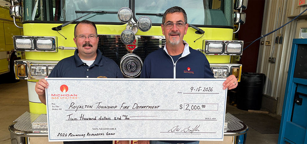 A man with MGU takes a photo with the Royalton Township fire chief while holding a ceremonial donation check in front of a fire engine in a garage.
